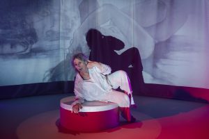 pia rosenbaumligger på sokkel med fotoprojektioner bag sig på en scene