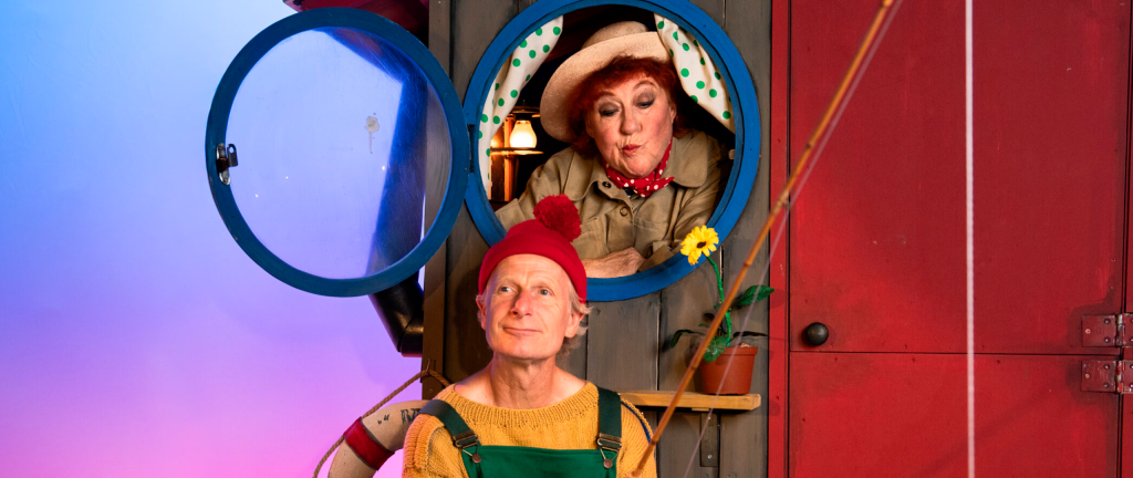 en kvinde kigger ud af et koøje i et hus og en mand sidder foran huset med en fiskestang